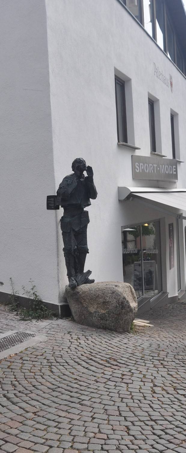 Gepflasterter Weg; links steht auf einem Stein eine lebensgroße schwarze Figur eines Mannes, der die Hände zum Mund hält, als würde er rufen.