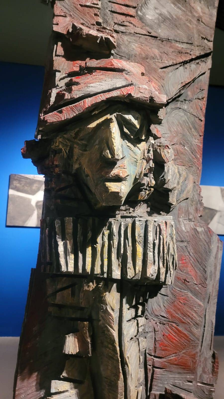 Detailaufnahme eines Holzkopfes mit überdimensionaler Halskrause und roter Mütze mit kantige Gesichtszüge, tiefe unregelmäßige Einschnitten mit roten Akzenten
