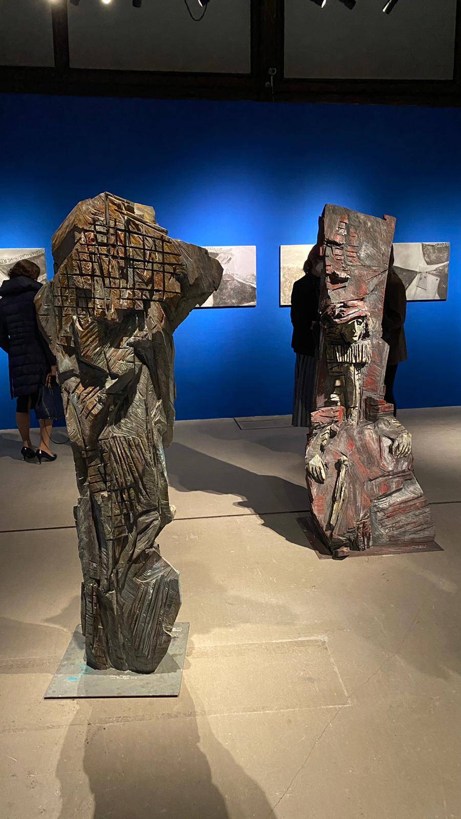 Lebensgroße Holzfigur auf Metallplatte sitzend mit Büchern und Zepter vor sich, sichtbaren Kettensägespuren und rauer, zerklüfteter Oberfläche in Rottönen, vor blauer Wand mit Alubildern