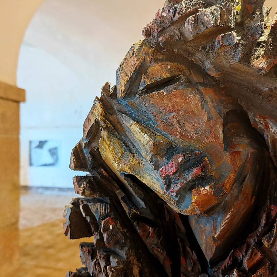 Detailaufnahme eines bunten Holzkopfes einer nach links geneigten Frau mit roten Lippen und tiefen Kettensägespuren vor hellem Hintergrund
