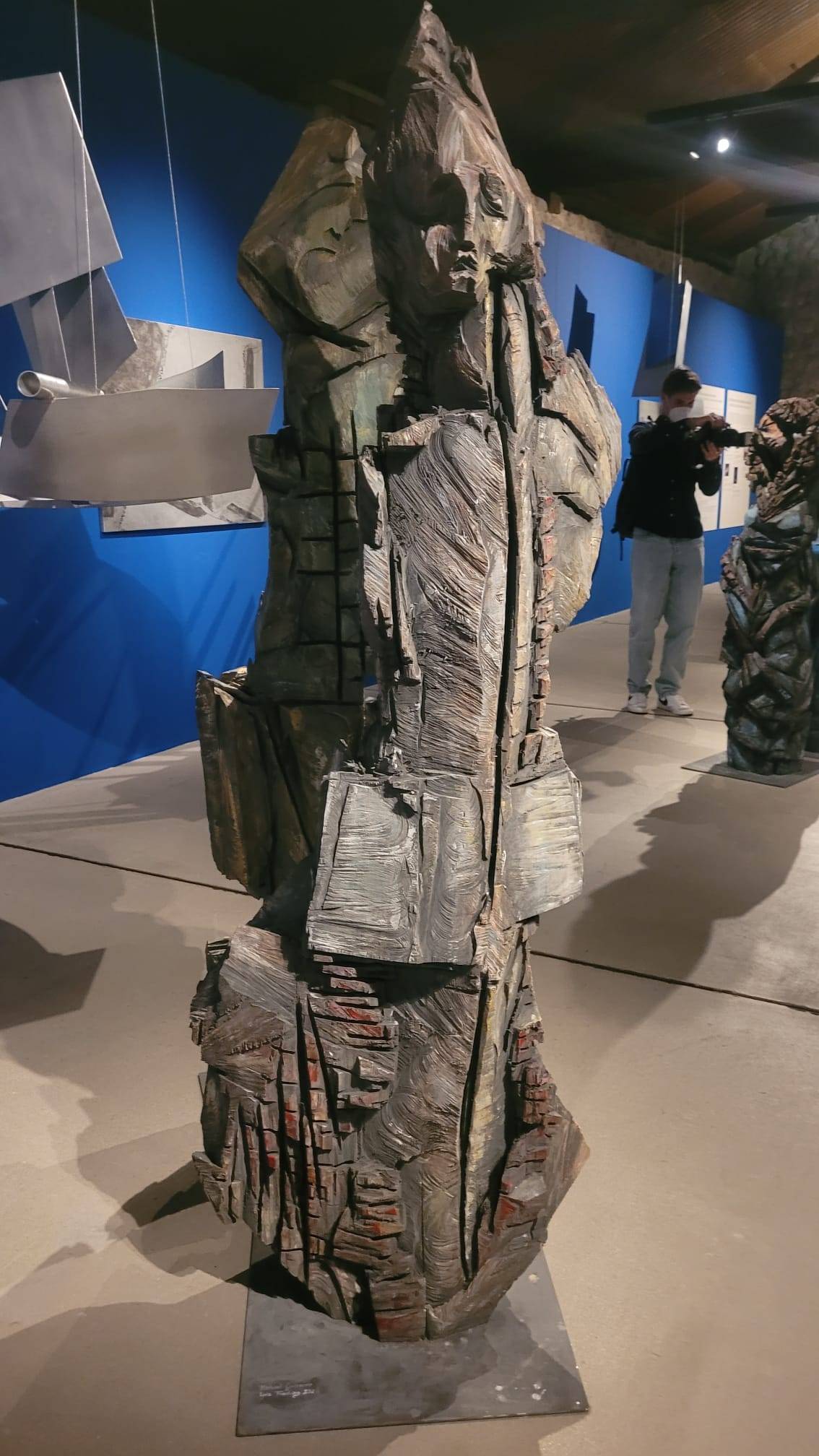 Lebensgroße graue Holzfigur auf Metallplatte mit tiefen Einschnitten, sichtbaren Kettensägespuren und roten Akzenten.